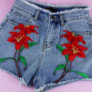 May include: Light blue denim shorts with frayed edges, adorned with embroidered red lily flowers and green leaves. The shorts have a classic five-pocket design and a button closure.
