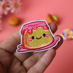 May include: A pink and green embroidered iron-on patch of a cartoon pudding with a happy face and pink frosting.