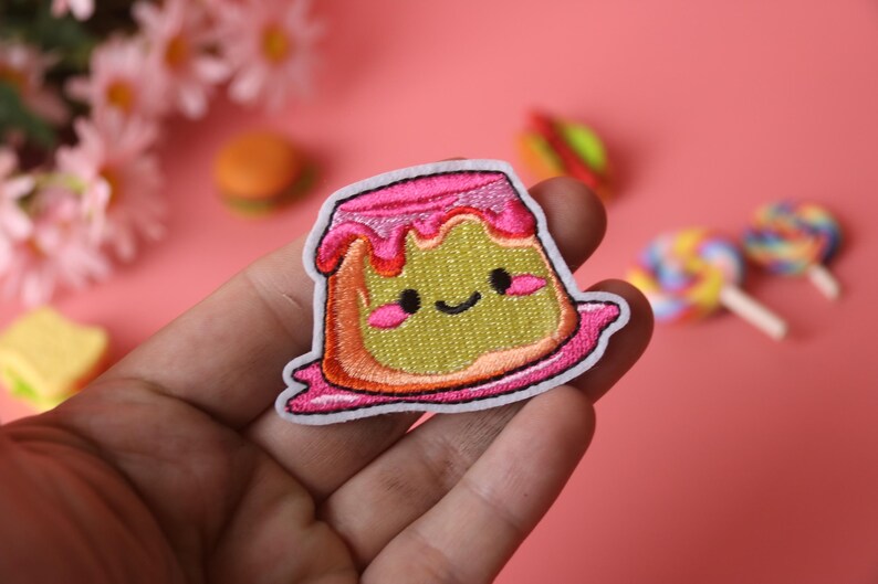 May include: A pink and green embroidered iron-on patch of a cartoon pudding with a happy face and pink frosting.
