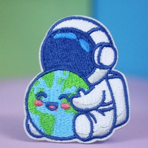 Op de afbeelding: Gestikte patch van een witte astronaut die een glimlachende blauwe en groene aarde omhelst. De astronaut heeft een blauwe helm en een wit ruimtepak.