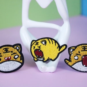 May include: Three embroidered iron-on patches featuring cartoon tigers with different expressions. The tigers are yellow with black stripes and have white accents.