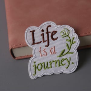 May include: Embroidered patch with the words "Life is a journey" in brown, pink, and green. The patch has a white border and a floral design. The patch is placed on a pink notebook.