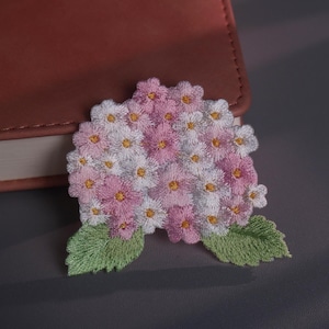 May include: Embroidered floral patch with pink and white flowers, yellow centres, and green leaves. The patch is on a grey surface beside a brown leather-bound book.