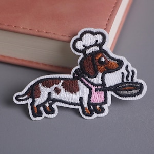 May include: Embroidered patch featuring a cartoon dachshund chef, wearing a chef's hat and pink apron, holding a frying pan. The dog is brown, white, and black, with steam rising from the pan. The patch is on a gray surface.