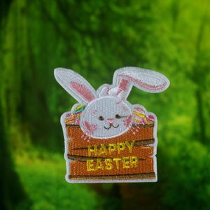 Könnte beinhalten: Ein weißer Hasenkopf schaut aus einer braunen Holzkiste mit dem Text "Happy Easter" in gelben Buchstaben.