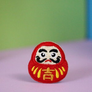 Puede incluir: Muñeco daruma rojo y amarillo bordado con una cara blanca y ojos y cejas negros. El muñeco tiene un bigote negro y una inscripción amarilla en la parte delantera.
