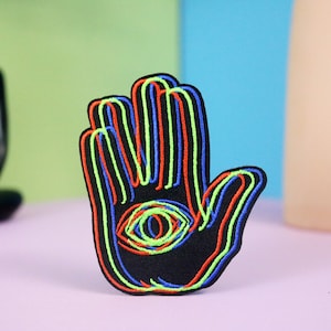 Könnte beinhalten: Ein schwarzer gestickter Aufnäher mit einer Hand mit einem Auge in der Handfläche. Die Hand ist mit rotem, blauem und grünem Faden umrandet.