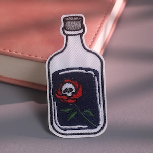 May include: An embroidered patch shaped like a glass bottle with a white outline and a dark blue interior. Inside the bottle is a red rose with a white skull at its center. The bottle has a grey cap at the top and a white border surrounding the design.