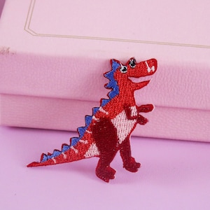 May include: Red and blue embroidered dinosaur patch. The dinosaur is standing on its hind legs with its mouth open and its tongue sticking out.