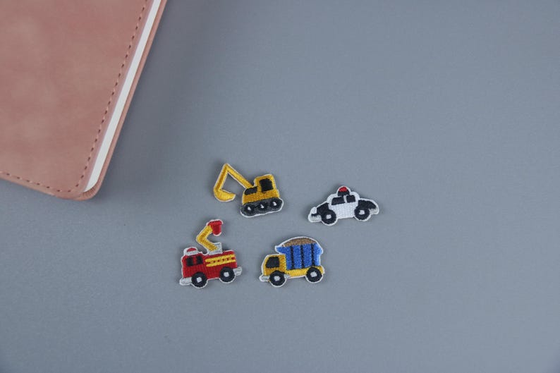 May include: Four embroidered iron-on patches featuring different vehicles: a yellow excavator, a red fire truck, a blue dump truck, and a white police car.