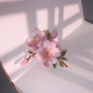 May include: Embroidered pink cherry blossom patch with green leaves and brown stems. The floral design is detailed with yellow accents in the center of the flowers. The patch is placed on a light pink surface.
