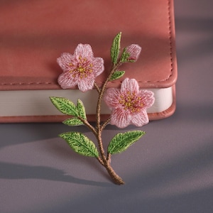 May include: Embroidered floral patch featuring pink cherry blossoms with yellow centers and green leaves. The patch is attached to a brown stem. The patch is placed on a pink leather-bound notebook.