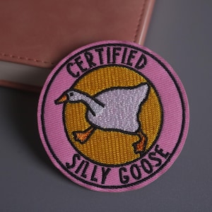 Op de afbeelding: Geborduurde patch met een roze rand en een gele kern met een cartoon gans. De patch leest "CERTIFIED SILLY GOOSE" in zwarte tekst. De gans is wit met oranje voeten en snavel.