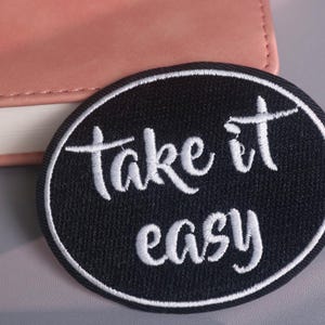 May include: Black oval embroidered patch with the words "take it easy" in white script. The patch has a white border and is placed on a gray surface next to a pink notebook.
