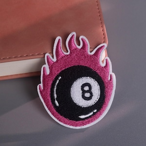May include: Embroidered patch featuring a black eight ball with the number 8 in white, surrounded by pink flames. The patch has a white border and is set against a pink notebook and gray surface. This patch is suitable for clothing and accessories.