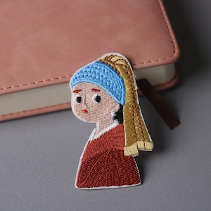 May include: Embroidered patch depicting a stylized portrait of a woman with a blue headscarf and a red dress. The patch is a colourful depiction of a famous artwork, ideal for adding to clothing or accessories.
