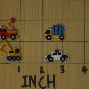 May include: Four embroidered iron-on patches featuring various vehicles: a red fire truck, a yellow excavator, a yellow dump truck, and a white police car. The patches are displayed on a wooden surface with a ruler marked in inches.