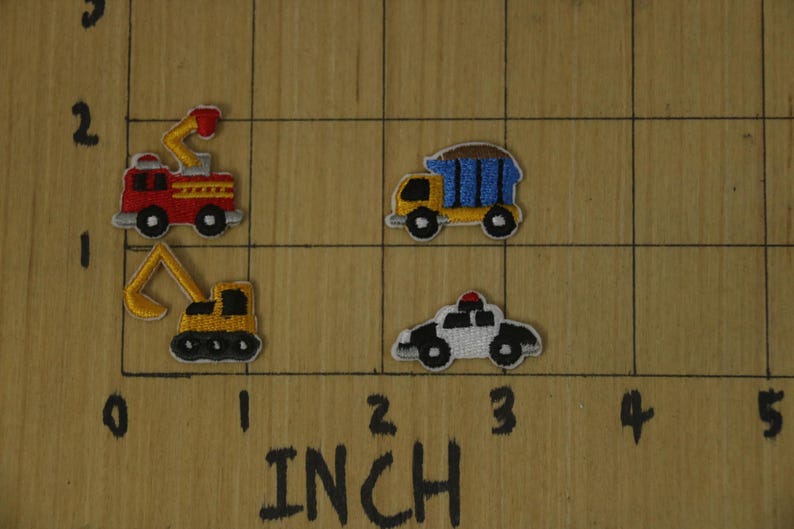 May include: Four embroidered iron-on patches featuring various vehicles: a red fire truck, a yellow excavator, a yellow dump truck, and a white police car. The patches are displayed on a wooden surface with a ruler marked in inches.