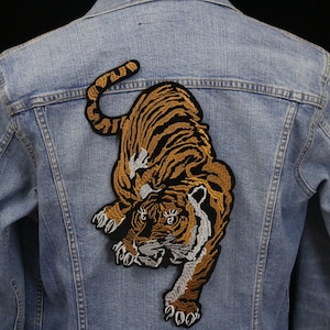 Può includere: Una giacca di jeans blu chiaro con una grande toppa di tigre ricamata sul retro. La tigre è arancione e nera con accenti bianchi.