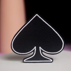 May include: A black spade-shaped patch with a white border. The patch is made of a textured fabric and is set against a blurred background with pink and black elements. The patch is a symbol often associated with playing cards.