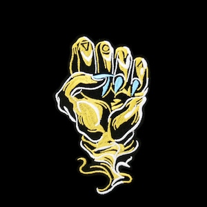 May include: A gold embroidered patch of a hand with long, sharp, blue fingernails. The hand is clenched into a fist and the background is black.