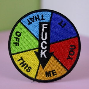 May include: Embroidered patch with a circular design featuring a segmented wheel with the word "FUCK" in the centre. The wheel is divided into sections with phrases like "OFF," "YOU," and "ME."