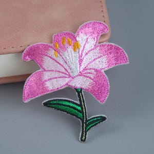 Pode incluir: Patch de flor bordada rosa com caule e folhas verdes. A flor tem um centro branco e detalhes amarelos.