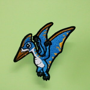 May include: A blue and gold embroidered iron-on patch of a cartoon pterodactyl with a white belly.