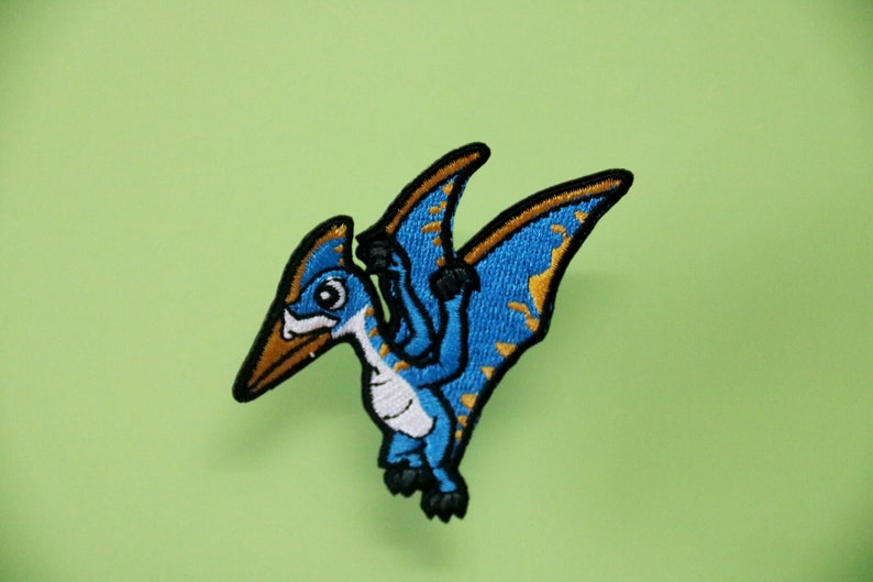 Pode incluir: Um remendo de ferro bordado azul e dourado de um pterod&aacute;ctilo de desenho animado com barriga branca.