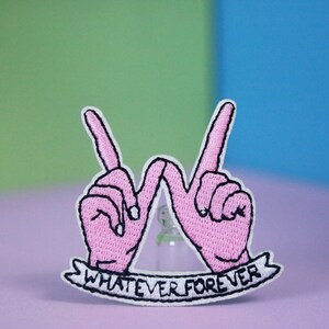 Könnte beinhalten: Rosa gestickter Patch mit zwei Händen, die ein "W" bilden, und einem Banner darunter, auf dem "WHATEVER FOREVER" steht.
