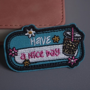 May include: Embroidered patch with a teal background and the words "HAVE A NICE DAY" in white and pink. A graphic of a bubble tea drink is on the right. The patch is decorated with flowers and a black border.