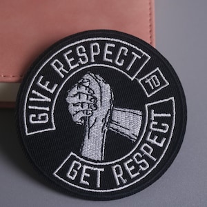 Può includere: Toppa ricamata in bianco e nero con la scritta "GIVE RESPECT TO GET RESPECT" in un design circolare. La toppa presenta un pugno chiuso al centro. La toppa è su una superficie grigia con un oggetto in pelle rosa sullo sfondo.