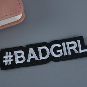 Pode incluir: Etiqueta bordada preta e branca com o texto "#BADGIRL" em letras brancas sobre fundo preto.