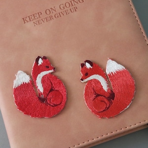 Könnte beinhalten: Zwei rote gestickte Fuchsflicken mit weißen Schwänzen, angebracht auf einem braunen Ledernotizbuch mit dem Text "Keep on going never give up".