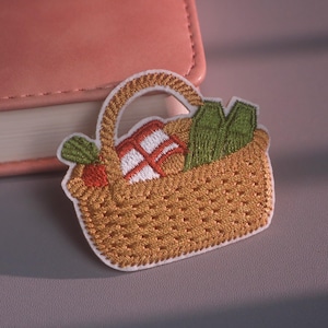 May include: An embroidered patch shaped like a woven picnic basket with a handle. The basket contains a red and white checkered cloth, a green leafy vegetable, and a small orange fruit, all outlined in white thread against a tan background.