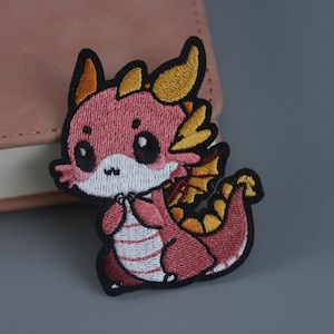 Pode incluir: Um remendo bordado de ferro de um dragão de desenho animado rosa e amarelo com uma expressão fofa.