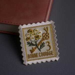 Puede incluir: Parche bordado con diseño de sello postal. El parche presenta un ramo de flores y una llave en tonos amarillos y dorados, con el número 3. También incluye el texto "BONNE COURAGE".