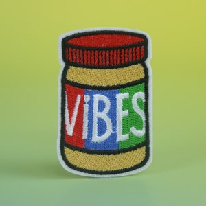 May include: A white embroidered patch shaped like a jar of peanut butter with the word "VIBES" written in red, blue, and green letters.