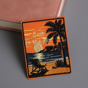 May include: Embroidered patch with a beach scene at sunset. The patch features a palm tree, a beach chair, and the text "Salt in the Air" against an orange and blue background. The patch has an orange border.