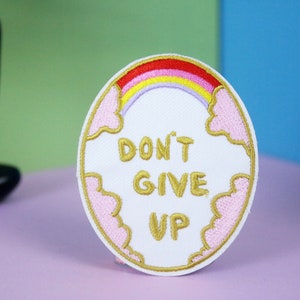 Könnte beinhalten: Weißer gestickter Aufnäher mit einem rosa Wolkenrand und einem Regenbogenbogen. Der Text "DON'T GIVE UP" ist in Gold gestickt.
