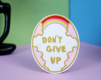 Könnte beinhalten: Weißer gestickter Aufnäher mit einem rosa Wolkenrand und einem Regenbogenbogen. Der Text "DON'T GIVE UP" ist in Gold gestickt.