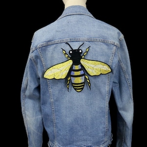 May include: Pale blue denim jacket with a large embroidered bee motif on the back. The bee design features yellow, black, and blue detailing. The jacket has a classic collar, button fastenings, and pockets.