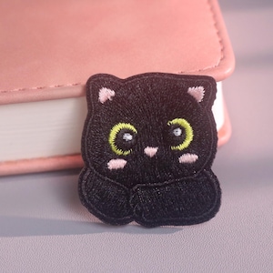 May include: An embroidered patch featuring a stylized black cat with large, bright yellow-green eyes, small pink inner ears, and light pink cheek accents. The cat is depicted with its paws tucked inward against its chest in a rounded, compact shape.