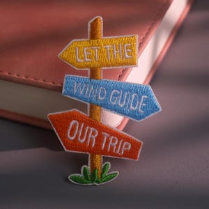 Op de afbeelding: Een geborduurde patch in de vorm van een wegwijzer met drie richtingspijlen. De bovenste gele pijl leest LET THE, de middelste blauwe pijl leest WIND GUIDE en de onderste oranje pijl leest OUR TRIP. Een klein groen grasveldje zit aan de onderkant.