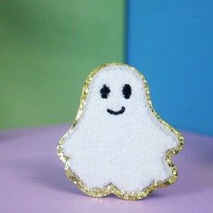 May include: A white ghost-shaped decoration with a gold glitter border. The ghost has a smiling face with black eyes and a mouth. The background is a mix of purple, green, and blue.