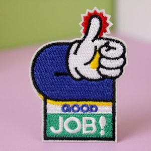 May include: Embroidered iron-on patch with a blue cartoon hand giving a thumbs up with a red star above it. The hand is holding a white sign with yellow trim that says "GOOD JOB!" in green letters.