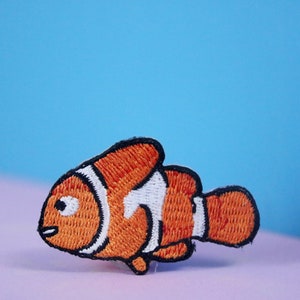 以下が含まれることがあります： オレンジと白の刺繍されたクマノミのパッチ。パッチは青とピンクの背景の上にあります。