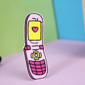 Pode incluir: Um remendo bordado para passar a ferro de um telemóvel flip rosa e amarelo com um coração no ecrã.