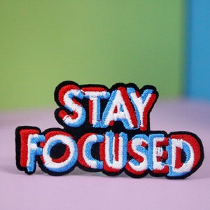 May include: A red, white, and blue embroidered patch with the text "STAY FOCUSED" in a bold, blocky font.