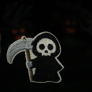 Peut inclure: Un patch brodé noir et blanc représentant un grim reaper de dessin animé tenant une faux. Le faucheur porte une robe à capuche et a un visage de crâne.